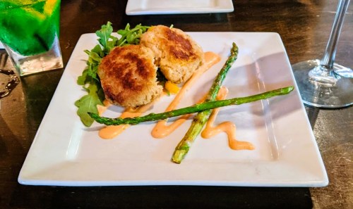 A plate with two golden crab cakes on greens, asparagus, and a drizzle of sauce, alongside a colorful drink.