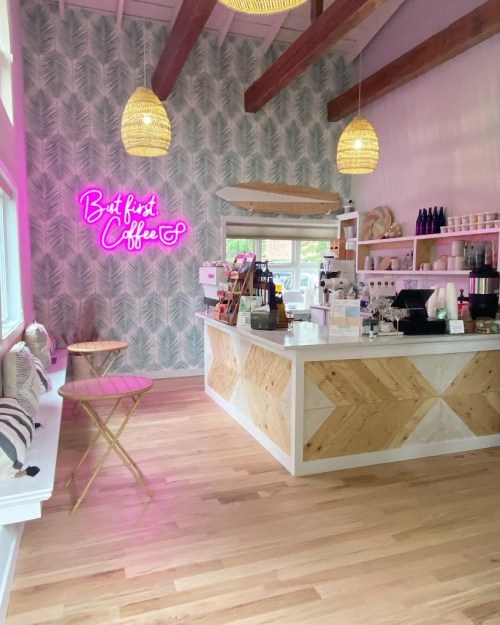 Bright coffee shop interior with pink neon sign, wooden counter, and stylish decor featuring palm leaf wallpaper.