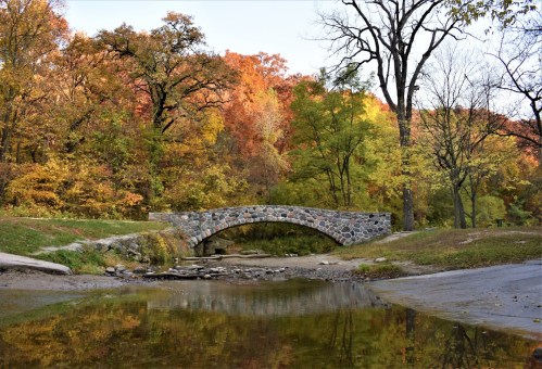 A stone bridge arches over a calm stream, surrounded by vibrant autumn foliage and trees.