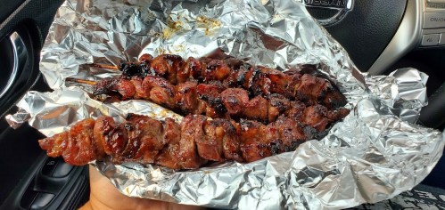Grilled skewers of meat wrapped in foil, held in a hand inside a car.