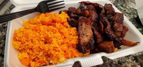 A takeout container with orange rice and grilled meat, served with a black fork on a granite surface.