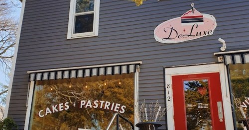 Exterior of DeLuxe bakery featuring a sign and large windows displaying cakes and pastries. Red door and striped awning.
