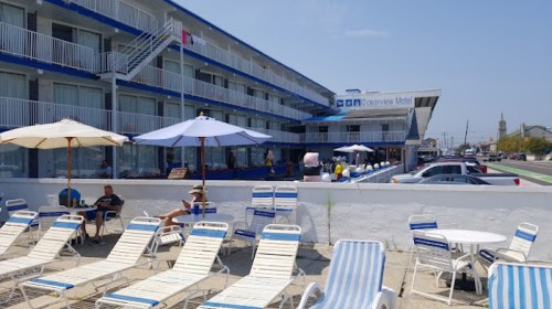 A beachfront motel with blue exterior, lounge chairs, umbrellas, and guests relaxing outside on a sunny day.