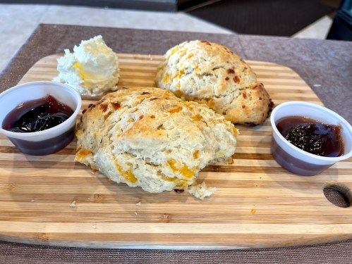 Two scones with orange bits on a wooden board, accompanied by butter and two small cups of jam.