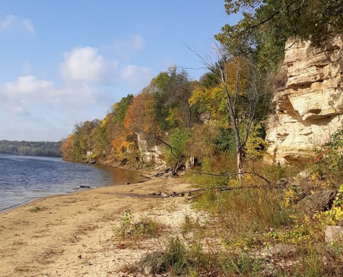 A sandy beach beside a calm river, with colorful autumn trees and rocky cliffs in the background under a blue sky.