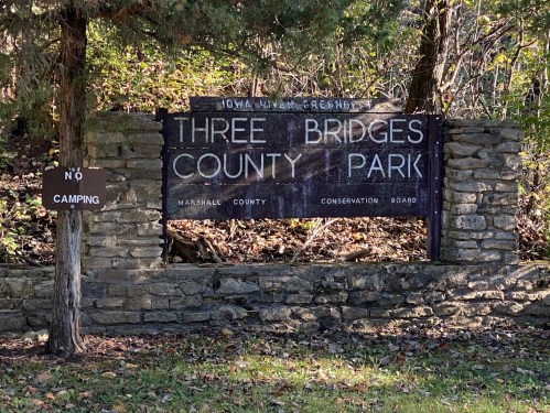 Sign for Three Bridges County Park, with a "No Camping" notice, surrounded by trees and stone structure.