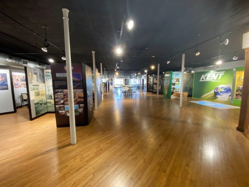 A spacious museum gallery with wooden floors, display panels, and bright lighting, showcasing various exhibits.
