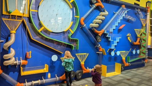 Two children interact with a colorful, interactive wall featuring pipes, funnels, and various mechanical elements.