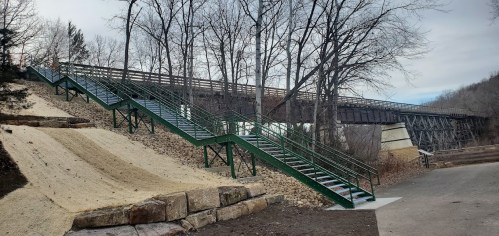 A green staircase leads up a slope, with a bridge visible above and trees lining the background.