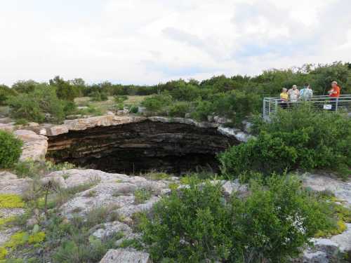 A group of people stands near the edge of a large sinkhole surrounded by greenery and rocky terrain.