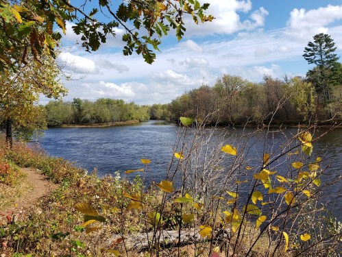 A serene river bends through a landscape of trees and autumn leaves under a partly cloudy sky.
