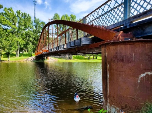 A rusted metal bridge arches over a calm lake, surrounded by green trees and a clear blue sky.