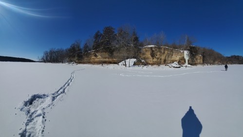 A snowy landscape with a frozen lake, a cliff in the background, and a person walking on the ice. Clear blue sky above.
