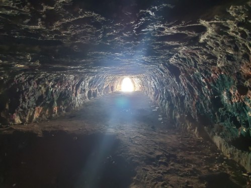 A dimly lit cave tunnel with colorful rock walls, leading to a bright light at the far end.