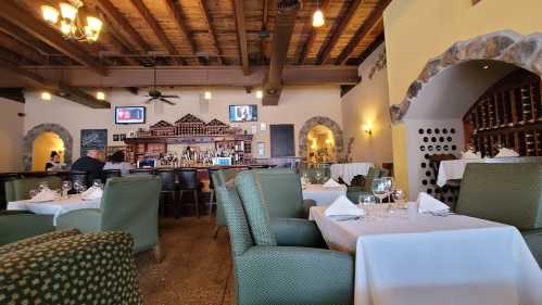 Interior of a restaurant featuring green chairs, white tablecloths, a bar, and rustic wooden beams.