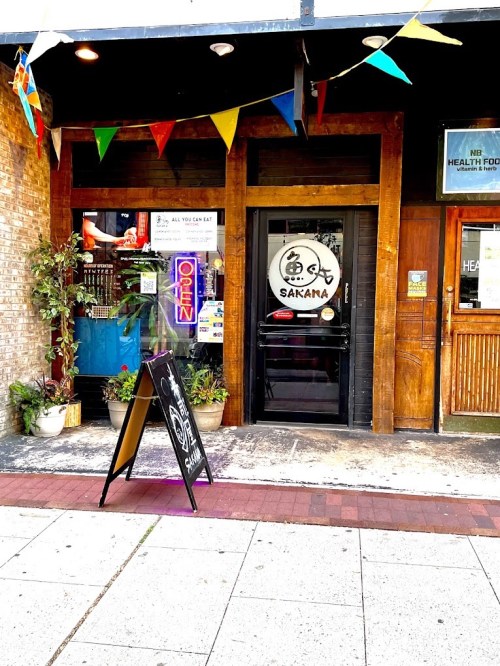 Exterior of Sakana restaurant with a wooden door, neon "Open" sign, and colorful flags above.