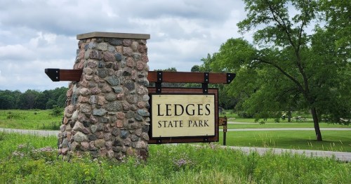 Sign for Ledges State Park, featuring a stone pillar and green landscape with trees and cloudy skies in the background.