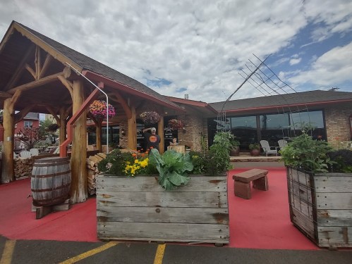 A rustic restaurant exterior with wooden features, colorful flower planters, and a red pathway under a cloudy sky.