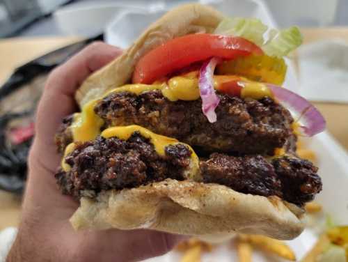 A hand holding a large burger with two patties, cheese, lettuce, tomato, and onion, alongside a side of fries.