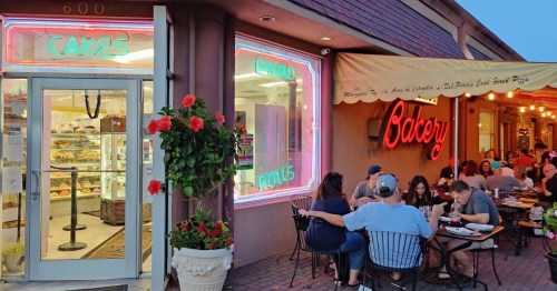 A bakery with neon signs, outdoor seating, and patrons enjoying food in a cozy atmosphere.