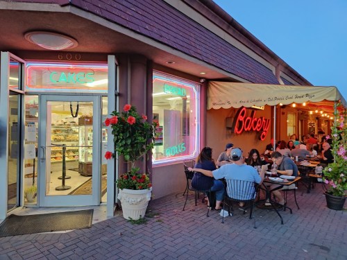 A bakery with neon signs, featuring outdoor seating where people enjoy food and drinks in a cozy atmosphere.