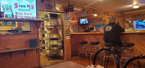 A cozy bar interior with wooden walls, a bartender, and a customer at the counter. A sign advertises drink specials.