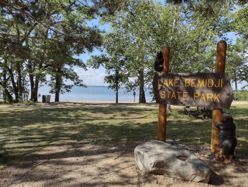Sign for Lake Bemidji State Park with trees and a view of the lake in the background.