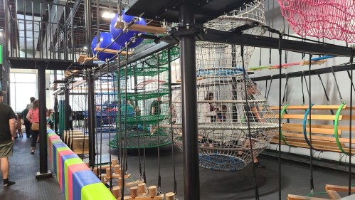 Indoor playground featuring climbing structures, nets, and colorful seating, with children and adults interacting.