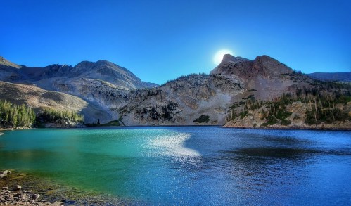 A serene mountain lake with turquoise water, surrounded by rocky hills and a clear blue sky, illuminated by the sun.