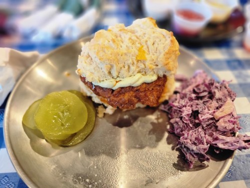 A plate with a biscuit sandwich filled with fried chicken, topped with sauce, and served with pickles and coleslaw.