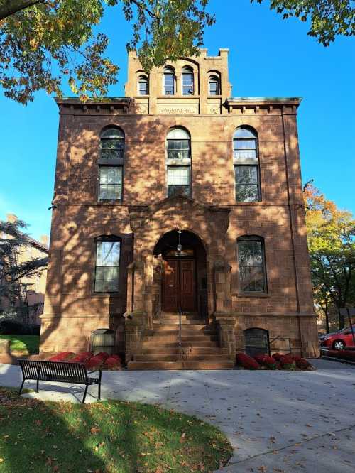 Historic brick building with arched entrance, large windows, and a tower, surrounded by trees and colorful foliage.