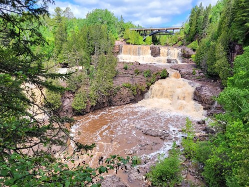 A scenic view of a waterfall surrounded by lush greenery and a bridge in the background under a partly cloudy sky.
