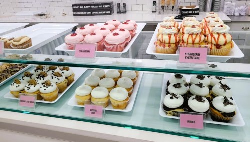 Display of various cupcakes, including strawberry, cheesecake, and marble, in a bakery case with labels.