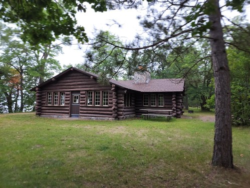 A rustic log cabin surrounded by trees, set in a grassy area near a lake.