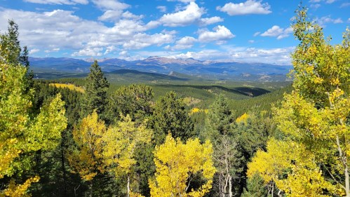 A scenic view of mountains and valleys, surrounded by vibrant autumn foliage under a blue sky with fluffy clouds.