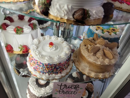 A display of colorful cakes, including a white frosted cake, a sprinkle-covered cake, and a peanut butter cake, with a sign.