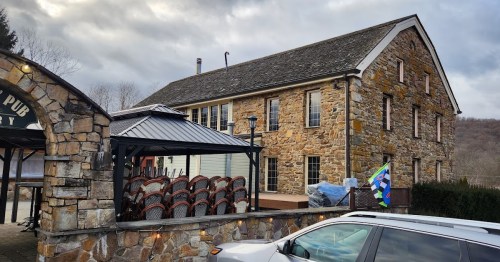 A rustic stone building with a sloped roof, featuring a patio area and stacked chairs outside.