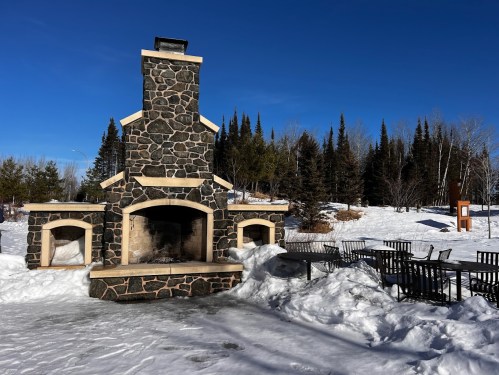 A stone outdoor fireplace surrounded by snow, with a seating area and trees in the background under a clear blue sky.