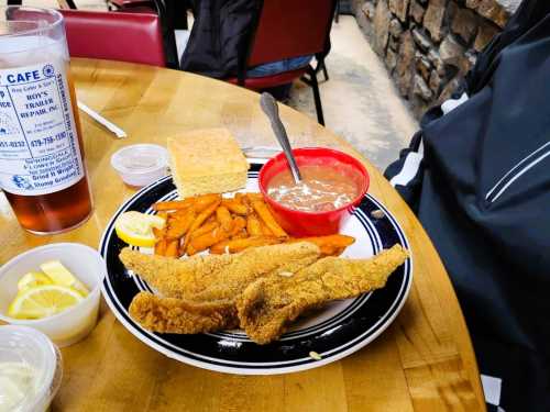 A plate of fried fish with sweet potato fries, cornbread, and a bowl of beans, served with lemon slices and iced tea.