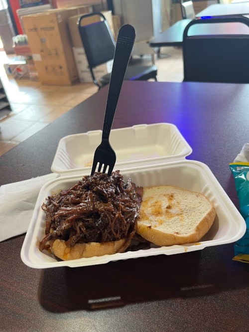 A pulled pork sandwich with a fork on top, served in a takeout container on a table.
