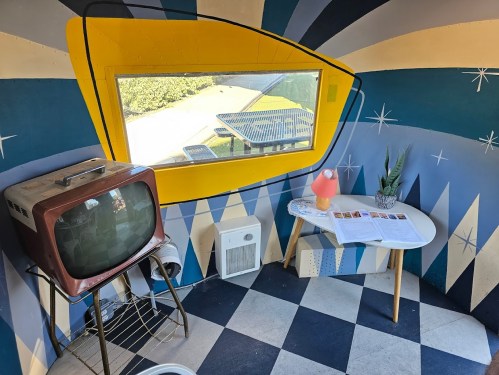 A retro room with a vintage TV, a small table, a lamp, and a large window featuring colorful geometric patterns.
