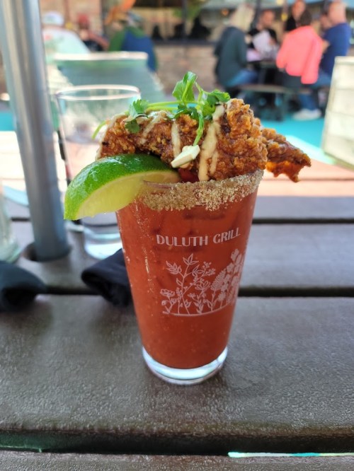 A glass of Bloody Mary garnished with a fried chicken piece, lime wedge, and herbs, sitting on a wooden table.