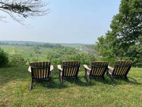 Four wooden lounge chairs facing a lush green landscape and river under a clear blue sky.