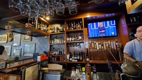 A bar with shelves of liquor, a TV displaying a show, and a bartender preparing drinks.
