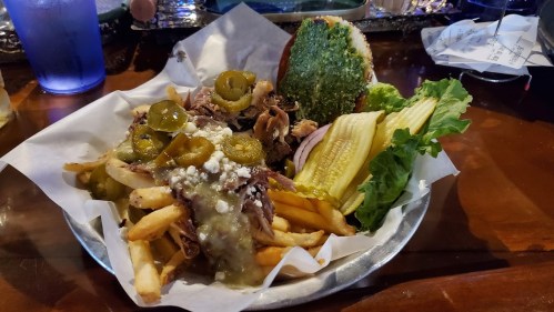 A plate of loaded fries topped with pulled pork, jalapeños, and cheese, served with pickles, lettuce, and a sandwich bun.