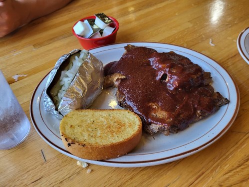 A plate with barbecue ribs, a baked potato, cornbread, and a small dish of condiments on a wooden table.
