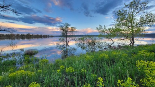 A serene lake at sunset, surrounded by lush greenery and trees, reflecting colorful clouds in the water.