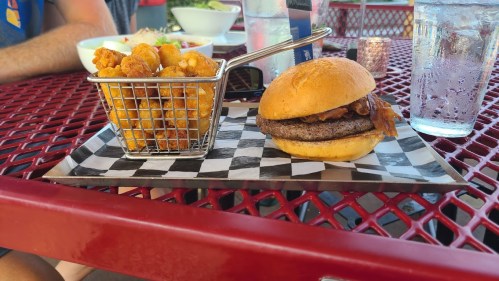 A burger with bacon on a plate next to a basket of crispy tater tots, set on a red outdoor table.