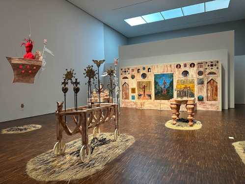 Art installation featuring wooden sculptures, vibrant paintings, and decorative elements in a spacious gallery.