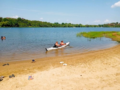 Two people in a kayak on a calm lake, with a sandy beach and greenery in the background.